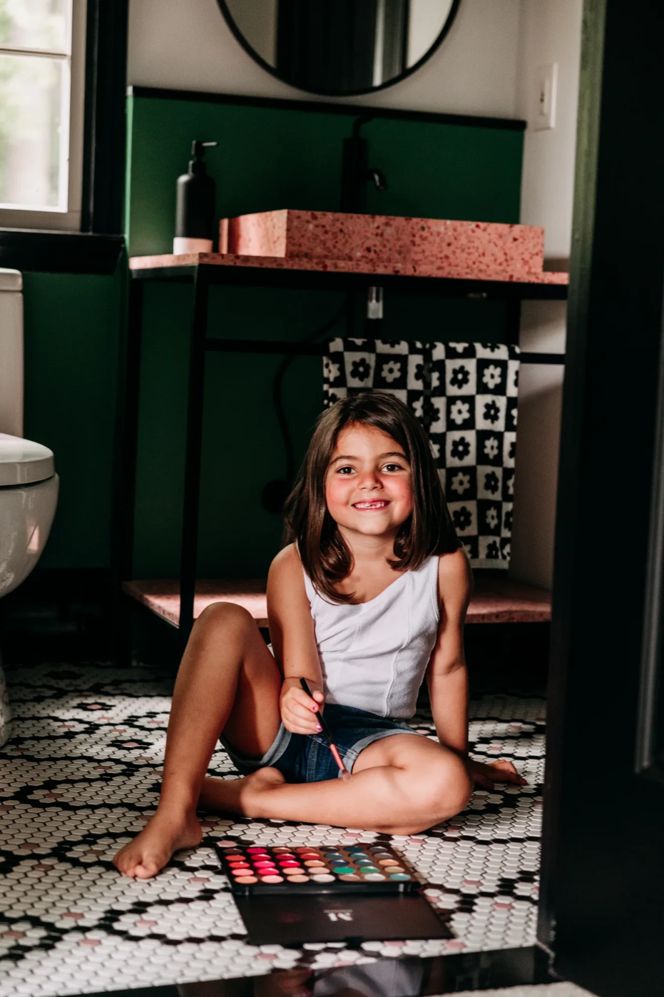 Child seated beside the console sink on the black, white, and pink mosaic floor.